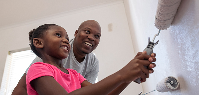 A young, smiling girl paints her new room with her father looking on.