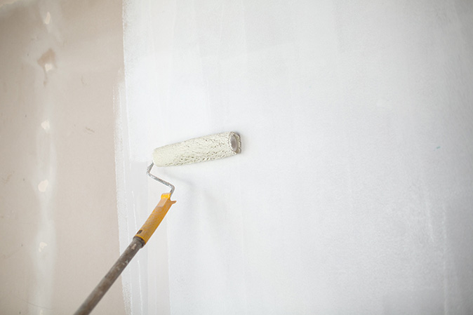 A person out of frame is holding a paint roller to apply white paint to freshly installed drywall.