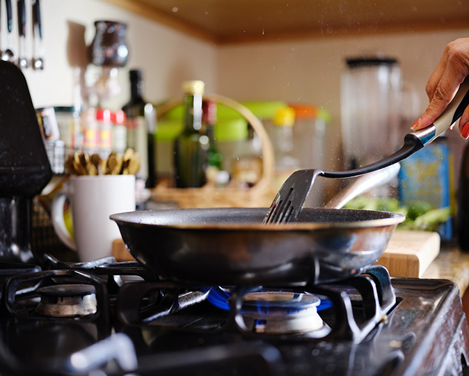 A homeowner cooks on a gas range using a stainless steel pan and a spatula.
