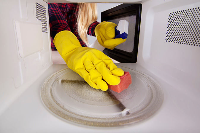 A woman wearing yellow gloves with a sponge and spray bottle cleaning a microwave.