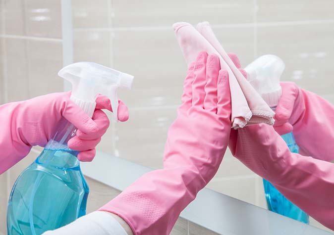 A person wearing pink rubber gloves is using the wrong cleaner in spray bottle to clean a mirror.
