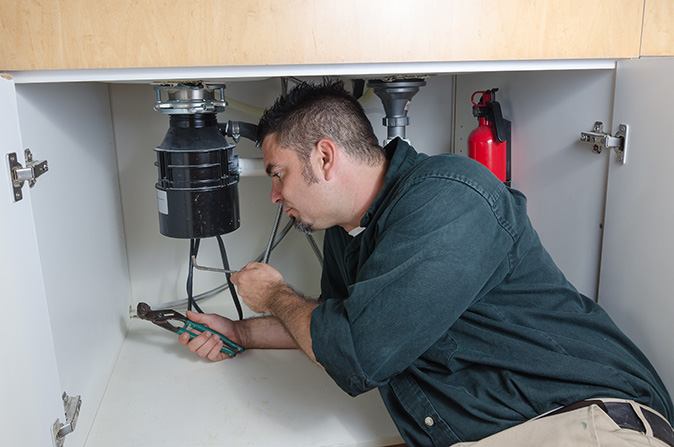A plumber is working under the kitchen sink to fix a broken garbage disposal.