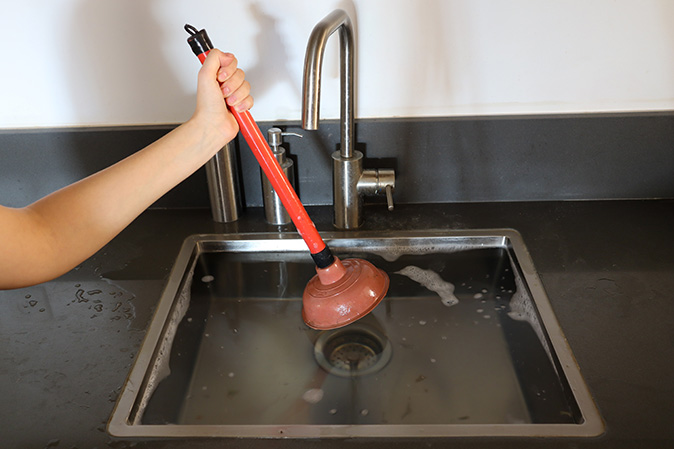 Person holding a plunger over a kitchen sink overflowing with water.