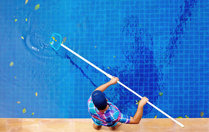 A young man using a pool skimmer to remove leaves and debris from the surface of a pool.