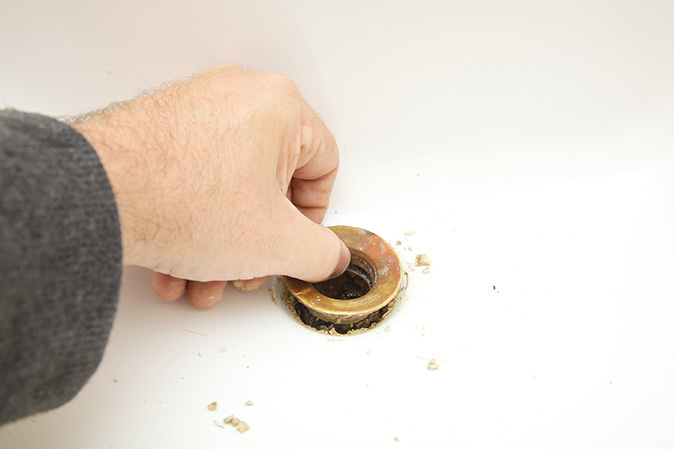 A man's hand removes a rusted drain ring from his bathroom sink to replace it with a pop-up stopper.