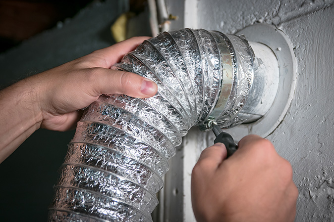 A close-up of a person’s hands using a screwdriver to unscrew a dryer vent hose from the wall.