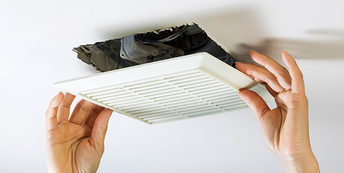A close-up of a homeowner holding up a new bathroom fan cover, showing the connectors in the housing next to the wiring coming from the wall.