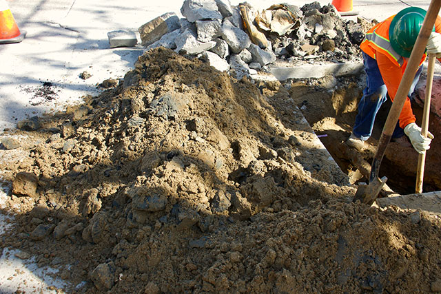 A member of a road crew stands in a hole made through concrete and dirt to fix a pipe.