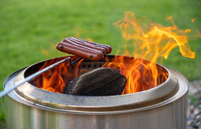 Closeup of hot dogs on a skewer being cooked over a stainless-steel smokeless fire pit.