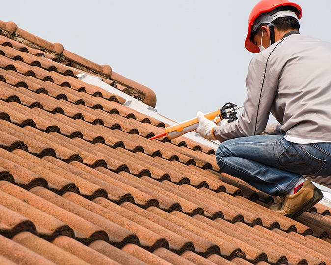 A roofer wearing protective gear replaces a damaged tile on a roof with a caulk gun.