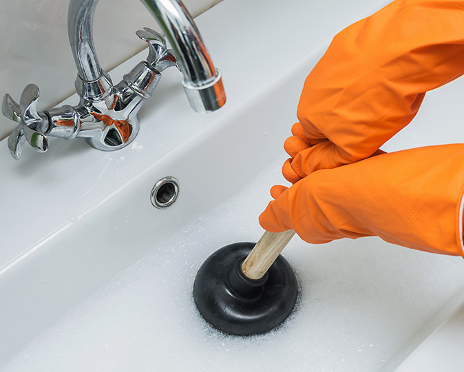 A person wearing orange rubber gloves uses a plunger to break up a drain clog.