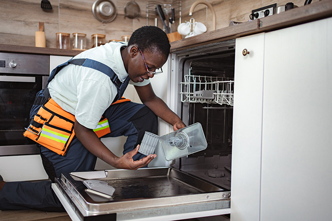 A man wearing an orange toolbelt inspects the dishwasher filter and housing in a dishwasher.