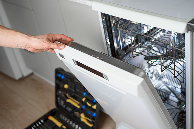 The hand of a repair technician opens the door of a dishwasher while a tool kit rests on the floor in the background.