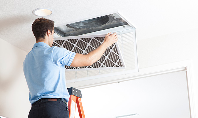 A technician standing on a ladder removes an air conditioning filter from an open vent.