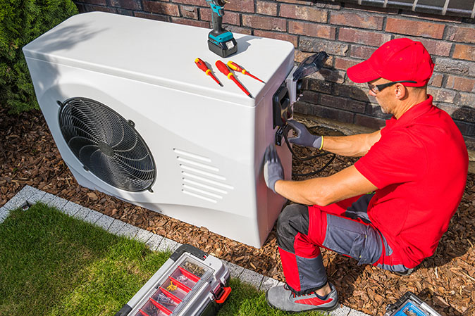 A repair technician with tools spread around inspects a backyard heat pump.