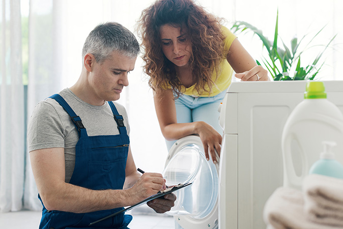 A repair technician writing on a clipboard after checking out a washing machine and showing the clipboard to a homeowner.