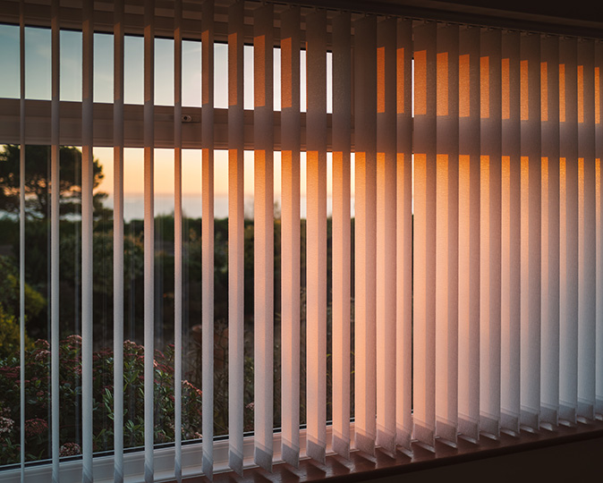 White vertical slat blinds hanging in front of a window as the sun is setting.