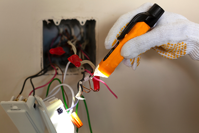 A close-up of a person’s hand replacing a light switch and using a voltage tester on the wires.