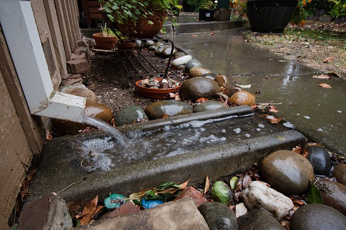 A downspout on a house with water pouring onto a concrete splash block, and over a sidewalk path during heavy rain.