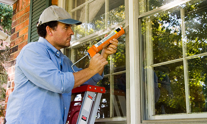 Person on ladder applies caulk to window in preparation for winter weather.
