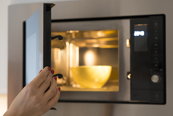 A woman opening the microwave to heat up soup in a yellow bowl.