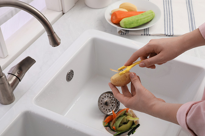 A woman is peeling potatoes into a kitchen sink before rinsing them into the garbage disposal for removal.