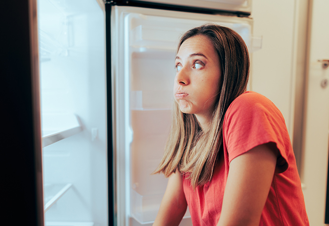 A Caucasian woman wearing a red shirt looks into an empty refrigerator with a frustrated expression